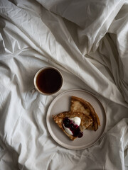 Breakfast in bed, a cup of black tea and thin pancakes with currant jam and sour cream on a beige ceramic plate on a soft white bed in the morning light