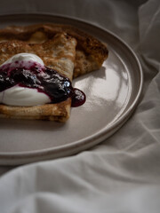 Breakfast in bed, a cup of black tea and thin pancakes with currant jam and sour cream on a beige ceramic plate on a soft white bed in the morning light