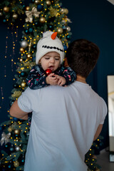 
baby in a snowman hat decorates the Christmas tree with dad