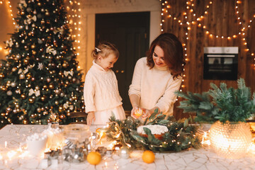 The concept of Christmas. Mom and little daughter child in cozy sweaters together prepare a festive cupcake meal in the decorated kitchen during the New Year holidays at home. Selective focus