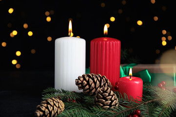 Burning candles, gift box, fir branches and cones on dark table against blurred Christmas lights