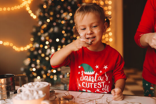 A joyful little girl child in red New Year's pajamas cooks, carves and eats ginger cookies in the decorated kitchen interior during the Christmas holiday at home. Selective focus