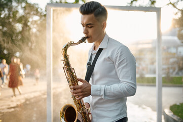Male saxophonist plays melody on saxophone in park - obrazy, fototapety, plakaty