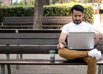Latin man using his laptop on a plaza bench with a coffee mug