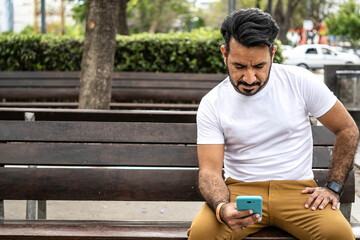 Latin man sitting on a plaza bench looking at his cell phone