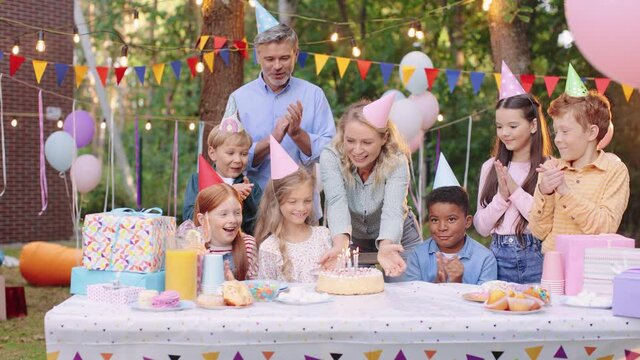 Happy children in party hats with party blowers at the birthday party applauding and having fun while mother bringing a cake