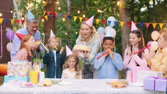 Happy children in party hats with party blowers at the birthday party applauding and having fun while mother bringing a cake