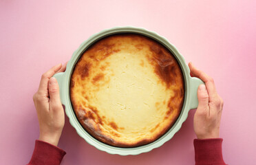 Cheesecake in baking pan with womans hands on pink background, from above overhead top view, flat lay, copy space, homemade food and bakery concept