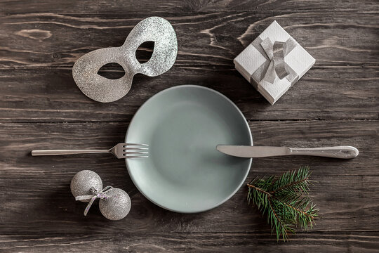 Festive table setting on a wooden table. New Year's decor: masquerade mask, gift box, Christmas balls, spruce twig and plate with knife and fork. Winter holiday concept. Place for text