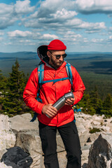 A traveler with a backpack on top of a mountain. Portrait of a traveler in a red cap and sunglasses. A tourist with a backpack and a thermos in his hands stands against the background of nature