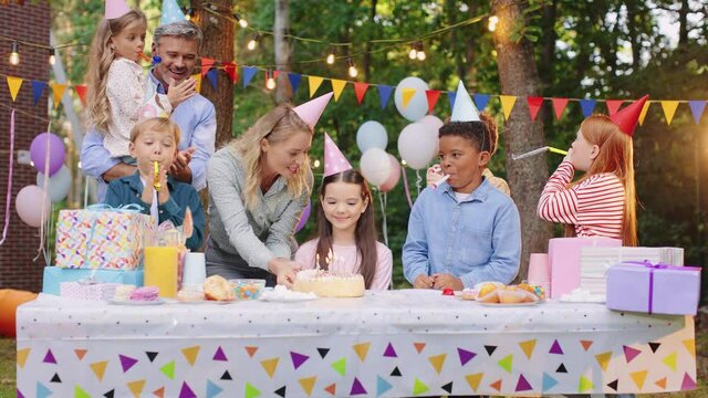 Caucasian cute birthday girl in conus sitting at the table with friends while her mother coming and bringing birthday cake. Holidays conceptCaucasian cute birthday girl in conus sitting at the table