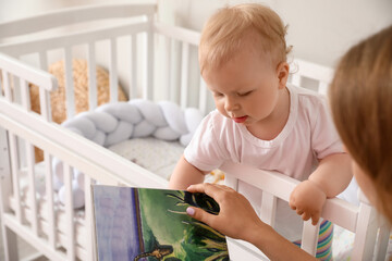 Mother reading book to her cute little baby in crib