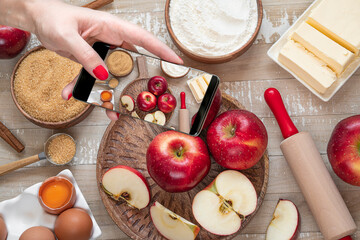 Female hand with mobile phone taking picture ofi for Apple pie or Charlotte on light wooden table. Ingredients for apple pie - red apples, flour, eggs, sugar, cinnamon and butter on a wooden table - Powered by Adobe