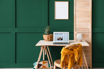 Modern workplace with laptop and blank frame hanging on green wall