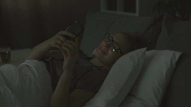 Positive mature lady with blond hair using modern smartphone before going to sleep. Caucasian woman wearing pajams and eyeglasses lying on comfy bed with soft linen.