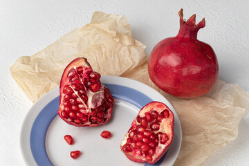 Still life with grenades. Pieces of fruit lie on a plate, next to a whole fruit on parchment paper. Horizontal orientation, white textured background, close-up.