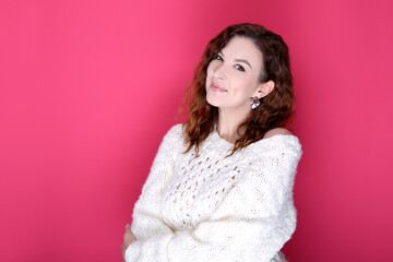 Smiling brunette woman girl in warm white sweater posing isolated on pink background studio portrait. Pretty girl looking at camera while standing with crossed arms.