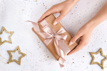 Female hands with natural manicure holding present in kraft paper with pink ribbon on white shiny background. Xmas composition. Flat lay. Happy holidays, New Year celebration and giving love concept. 
