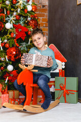 Obraz na płótnie Canvas Little boy with christmas gift box in hands riding wooden toy horse under Christmas tree with gifts