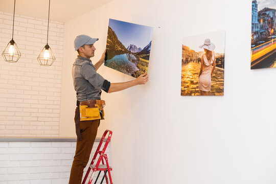 Young man standing on stepladder while his wife helping him to hang picture on wall in living-room of new flat or house