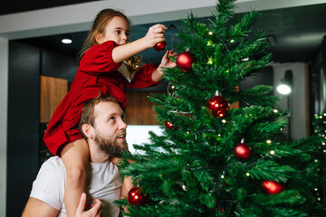 Cute little girl sitting on her dad's shoulders decorating a christmas tree with red shiny balls....