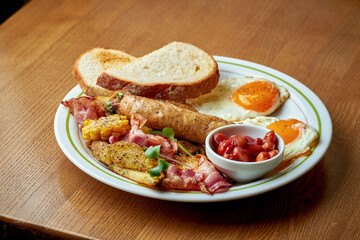 English breakfast - scrambled eggs with bacon, beans, sausage in a white plate on a wooden background