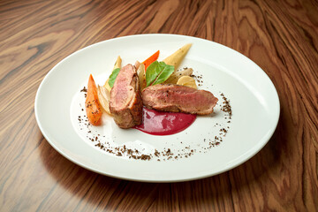 Duck fillet with baked vegetables and red sauce in a white plate on a wooden background