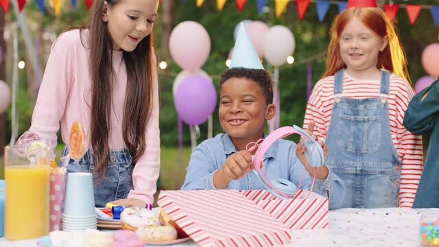 Portrait of multiracial child boy opening present and taking headphones at the table with his friends. Surprise present, birthday gift, happy kids, childhood concept