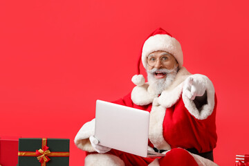 Santa Claus with laptop pointing at viewer on color background