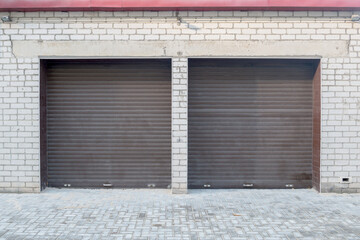 Double garage is made of white brick with brown folding doors like blinds with lower lock, two lighting lamps and red visor, air vent at bottom, on ground  area paved with gray street tiles.