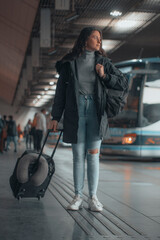 Viajera de empresa de cabello moreno observando impaciente de pie esperando con su mochila y maleta de viaje en la estación de autobuses por el recinto para tomar su trayecto de transporte