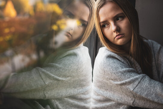 Depressed and anxious young woman sitting by a large window, feeling blue, sad, uncertain