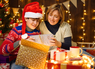 Fototapeta na wymiar Portrait of mother and son in new year decoration. Woman and teen boy opening the box, take out gifts, having fun and laughing. Holiday lights, gifts and christmas tree decorated with toys.