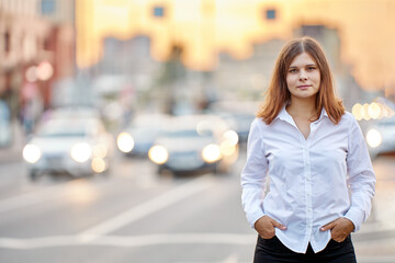 Fototapeta premium Woman with her hands in her pockets stands on city street with heavy traffic in summer at sunset.