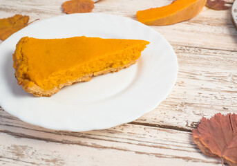A slice of American pumpkin pie and pumpkin on a wooden white table. Traditional fall food for Halloween, Thanksgiving.