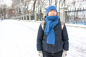 Young European woman 30-40 years old looking smiling into camera standing in snow-covered winter park. A road going into perspective. Girl wearing blue scarf hat with backpack in cold day. Copy space.