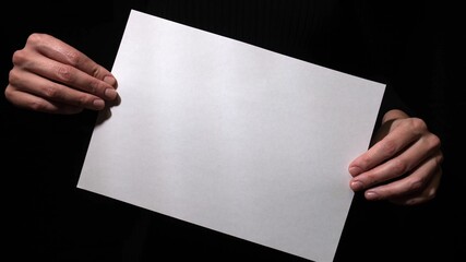 Hands holding blank white empty sheet of paper on black background, suitable for text and signs