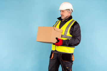 Builder is holding a closed carton box. Worker with a box in his hands. Concept - it works in warehouse. Portrait of a warehouse worker in helmet and uniform. Warehouse worker on turquoise background