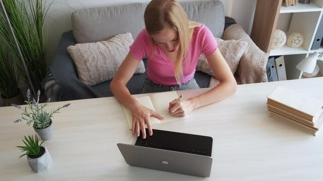 Distance education. Female student. Home learning. Concentrated woman writing in workbook with laptop on table in light room interior.