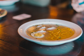 Side view of dietary soup with chicken meat balls, egg and noodles in white plate stands on the table. Pasta. Greens. Meal
