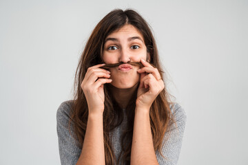 Portrait view of the adorable funny girl with brunette hair fooling around, making ridiculous childish comical grimace and playing with her hair. Indoor studio shot isolated on white background