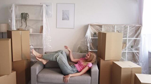 New home. Happy woman. House moving. Pretty lady moving sofa laying on with many carton boxes covering property in light room interior.