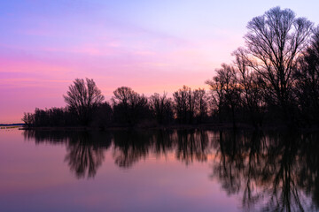 Orange and pink sunset over the lake with silhouettes of trees without leaves in the background 