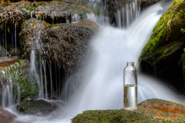 A half-full bottle of drinking water stands on a stone in front of a waterfall. By the middle of...