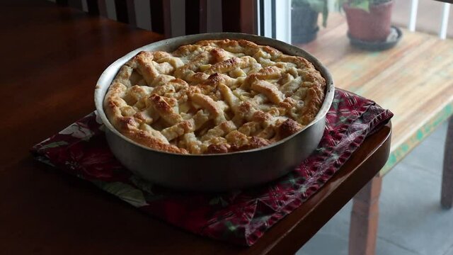 Female hands puts hot and homemade apple pie to rest on top of a wooden table with red cloth.