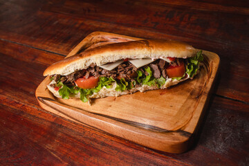 Long sandwich with meat, tomato and lettuce on a board on a wooden table.