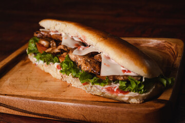Approach of a long sandwich of grilled chicken, tomato and lettuce on top of a board on a wooden table.