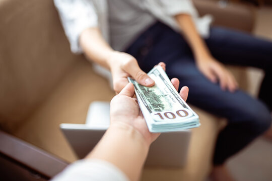 Mother Gives Her Son A Bundle Of Dollars. The Client Pays For The Work By Handing Over Money. Woman's Hand Passing A Bundle Of Dollars To A Man