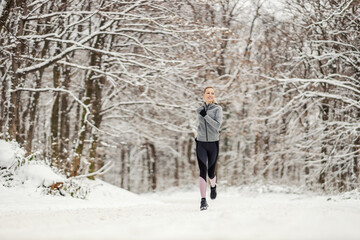 Happy fit sportswoman jogging in nature on snowy path at winter. Healthy habits, winter fitness, cardio exercises