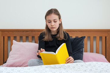 Young school age girl sitting on bed in bedroom and reading from yellow textbook. School homework.